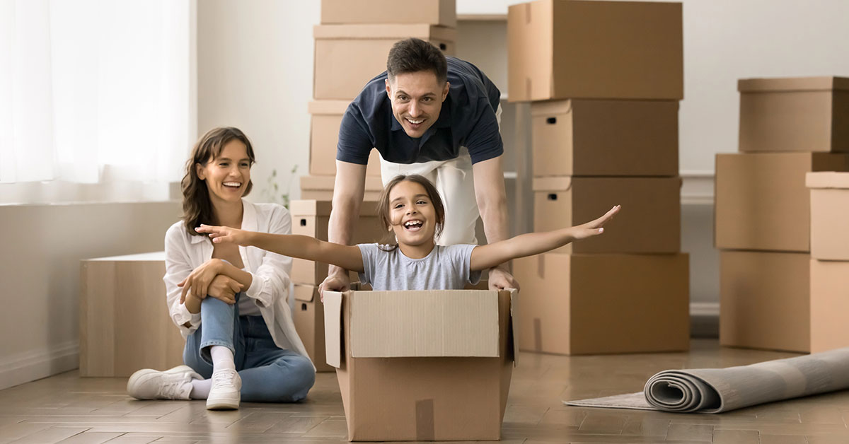 Glückliche Familie mit Eltern und Kind zwischen Umzugskartons, die gemeinsam den Einzug ins eigene Zuhause feiern – Symbol für Immobilienkauf und staatliche Förderung für Familien. | Immobilienfinanzierung